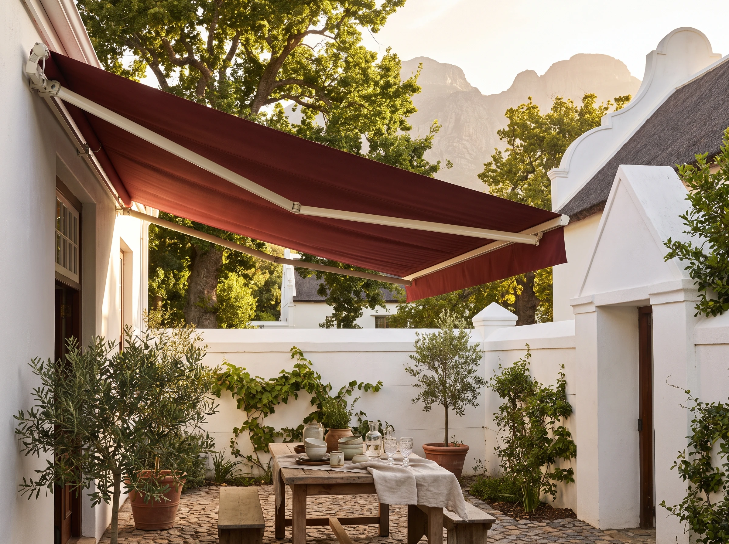 A Franschhoek gallery courtyard with a bordeaux folding-arm awning extended over a set table under oak trees